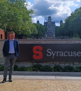 Dr Michał Krotoszyński na tle budynku Syracuse University College of Law i znaku "Syracuse"